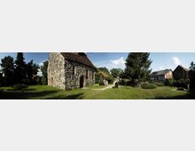 Steinkirche mit grob behauenem Mauerwerk und rotem Ziegeldach, umgeben von einer Grünfläche und Bäumen; neben der Kirche stehen weitere Gebäude im Dorfstil sowie ein kleiner Friedhof.