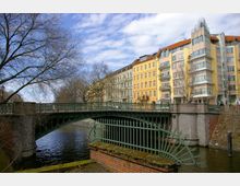 Blick auf eine grüne Metallbrücke über einen Fluss mit angrenzenden gelben und hellen Wohngebäuden im Hintergrund. Im Vordergrund sind ein dekorativer Zaun und ein gemauerter Sockel zu sehen, umgeben von kahlen Baumästen.