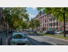 Straßenszene in einer Stadt mit breiter Fahrbahn, parkenden Autos und Bäumen, die Schatten spenden. Auf der rechten Seite steht ein großes Backsteingebäude mit grünen Markisen, links sind bunte Fassaden und Gehwege mit Fußgängern zu sehen.