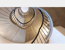 Ansicht einer Wendeltreppe von unten, mit Betonstufen und einem Geländer aus schwarzen Metallstreben, die spiralförmig nach oben führen. Die Umgebung ist schlicht, mit hellen Wänden und sichtbarer Betonstruktur.