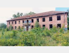 Verfallenes zweistöckiges Ziegelgebäude mit zerstörten Fenstern, Graffiti an den Wänden und umgeben von dichter Vegetation. Das Gebäude befindet sich in einer überwucherten, naturbelassenen Umgebung mit Bäumen und hohen Gräsern.