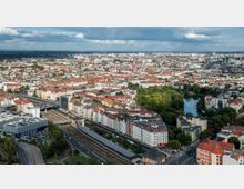 Luftaufnahme einer städtischen Landschaft mit dicht bebauten Wohnvierteln, zahlreichen Gebäuden mit roten Dächern und einem Park mit einem See im Hintergrund. Im Vordergrund verlaufen Bahngleise und Straßen mit Brücken und angrenzenden modernen Gebäuden.