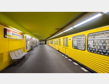 U-Bahnhof mit gelb gestrichenen Wänden und Decken, einem gelben U-Bahnzug der Linie U2, der am Bahnsteig "Bismarckstraße" hält, sowie Sitzbänken und Hinweisschildern an der Wand. Beleuchtungslampen ziehen sich entlang der Decke, und der Boden ist dunkelgrau.