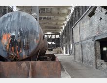 Innenansicht einer verlassenen Industriehalle mit hohen Decken und großen Fenstern, die teilweise beschädigt sind. Im Vordergrund steht ein rostiger, großer Tank mit einem Graffiti, umgeben von heruntergekommenen Backsteinwänden und Trümmern.