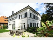 Weiße zweigeschossige Villa im klassischen Stil mit schwarzen Fensterläden und großen Fenstern, umgeben von einem gepflegten Garten. Im Vordergrund befindet sich eine gepflasterte Terrasse mit einem Holztisch und Stühlen.