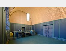 Innenraum eines Gebäudes mit hoher gewölbter Decke, ausgekleidet mit einem orange-weißen Rautenmuster. Die Wände sind dunkelblau mit grünen Zierelementen, und es gibt einen erhöhten Empfangsschalter sowie eine große, ornamentierte Holztür auf der rechten Seite.