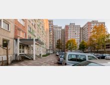 Eine Wohnanlage mit mehreren Plattenbauten, die im Stil des sozialen Wohnungsbaus gestaltet sind. Im Vordergrund befindet sich ein Parkplatz mit geparkten Autos, während rechts und links von Bäumen gesäumte Gehwege sichtbar sind.