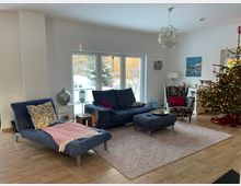 Helles Wohnzimmer mit großen Glas-Schiebetüren zur verschneiten Terrasse, Holzfußboden und hellem Teppich. Im Raum stehen ein dunkelblaues Sofa mit Hocker, eine Liege, ein gemusterter Sessel, ein Sideboard sowie ein geschmückter Weihnachtsbaum und moderne Deckenleuchte.