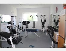 Kleiner Fitnessraum mit mehreren Fitnessgeräten, darunter Hantelbänke, eine Latzugmaschine und Hanteln, die ordentlich neben einer Spiegelwand gestapelt sind. Das helle Zimmer hat weiße Wände, einen grauen Boden und ein Fenster, durch das Tageslicht einfällt.