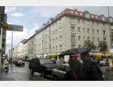 Straßenszene mit mehrstöckigen Wohnhäusern mit roten Ziegeldächern und Geschäften im Erdgeschoss. Im Vordergrund sind parkende Autos und Fußgänger unter einem Regenschirm zu sehen, während ein nasses Kopfsteinpflaster auf kürzlichen Regen hindeutet.