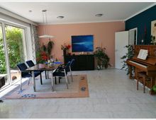 Wohnzimmer mit glänzendem weißen Marmorboden, einem Esstisch mit blauen Stühlen auf einem beigefarbenen Teppich und vielen Pflanzen. Im Hintergrund sind eine rosa Wand mit einem abstrakten Gemälde, ein Klavier neben einer dunkelblauen Wand und eine große Fensterfront zu sehen.