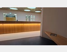 Moderner Innenraum mit langer, indirekt beleuchteter Holztheke vor einer weißen Wand mit großen Fensteröffnungen und Tür. An der Decke sind runde Lichtfelder in einer perforierten Deckenverkleidung, rechts befindet sich eine schmale Holzbank an der Wand.