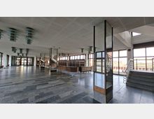 Weitläufige Eingangshalle eines öffentlichen Gebäudes mit grauem Steinboden, abgehängter Decke mit runden Leuchten und großen Fensterfronten. Rechts führen breite Treppen mit Metallgeländern zu einem erhöhten Bereich; im Hintergrund sind Glastüren und ein Kassen- oder Tresenbereich sichtbar.