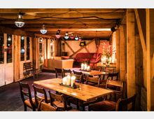 Gemütlicher Innenraum mit Holzbalken, Holztischen und Stühlen, dekoriert mit brennenden Kerzen. Im Hintergrund befinden sich Polstermöbel, Fenster mit Holztüren und ein Blumenstrauß auf einem Seitentisch.