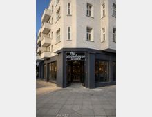 Ecke eines mehrstöckigen Wohn- und Geschäftshauses mit heller Putzfassade und Balkonen; im dunklen Erdgeschoss befindet sich ein Restaurant mit großen Schaufenstern und einem Eingang unter dem Schild „the smokehouse“. Vor dem Gebäude liegt ein breiter Gehweg mit Pflaster- und Steinplattenbelag.