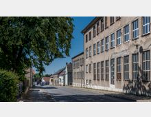 Straßenszene in einem Wohngebiet mit einer Reihe von älteren Gebäuden, darunter ein großes, verputztes Gebäude mit zahlreichen rechteckigen Fenstern rechts im Bild. Links steht ein großer Baum, der teilweise die Straße und gegenüberliegende Häuser beschattet.