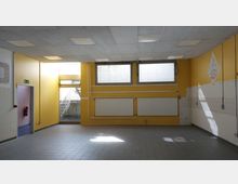 Großer leerer Raum mit gelben und weißen Wänden, rechteckigen Fenstern im oberen Bereich und grauem Fliesenboden. An den Wänden sind Heizkörper, sichtbare Rohrleitungen und Fliesenteile angebracht; im Hintergrund führt eine Tür zu einer Außentreppe.