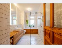 Modernes Badezimmer mit weißen Fliesen an Wänden und Boden, einer Holz-Waschtischplatte mit eingelassenem Waschbecken, einer Dusche mit Glasabtrennung und einer Badewanne mit Holzverkleidung unter einem großen Fenster. Pflanzen und eine Holztür ergänzen die Einrichtung.