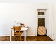 Ein heller Raum mit minimalistischem Schreibtisch, Stuhl und Schreibtischlampe vor einer weißen Wand. Rechts im Bild befindet sich ein dekorativer Kamin oder Kachelofen mit weißen Kacheln und Messingtüren.