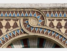 Detailansicht eines dekorativen Fassadenelements aus gemusterten Ziegeln, mit einer Mischung aus braunen, beigen und blauen Ornamenten. Das Design zeigt bogenförmige Linien, florale Motive und geometrische Muster unterhalb einer weißen Ziegelwand.