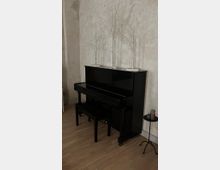 Ein schwarzes Klavier mit zugehöriger Bank steht vor einer hellen, strukturierten Wand. Auf dem Klavier stehen drei weiße, dekorative Bäume, daneben ein kleiner, schwarzer Beistelltisch mit Kerzen.