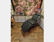 Innenraum eines Tattoo- oder Kosmetikstudios mit einer schwarzen, verstellbaren Behandlungsliege auf Rollen vor einer langen Arbeitsfläche mit vielen Schubladen. An der Rückwand hängt eine großflächige Tapete mit rosa Blumenmotiv; auf der Ablage stehen kleine Fläschchen, Utensilien und gerahmte Bilder.