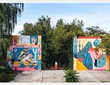 Bunte Außenwand mit großflächigem Streetart-Graffiti, bestehend aus abstrakten und urbanen Motiven, links eine Figur mit grafischen Symbolen, rechts eine Skyline und geometrische Formen; die Wand steht auf einer betonierten Fläche und ist von Bäumen und Sträuchern umgeben. Eine Person in weißem Oberteil und roter Hose steht zwischen den bemalten Wänden.