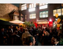 Großer Innenhof vor einem Ziegelgebäude mit leuchtendem Schild „Ballhaus“, umgeben von Pflanzen und Schirmen. Eine Menschenmenge steht zusammen, während Luftballons und Beleuchtung eine festliche Atmosphäre schaffen.