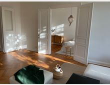 Wohnraum in einem hellen Altbau mit weißen Wänden und Holzdielenboden. Durch geöffnete Flügeltüren ist ein Schlafzimmer mit minimalistisch eingerichtetem Bett, Nachttisch und einer hängenden Korblampe zu sehen.