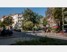 Ruhige Straßenkreuzung in einem Wohnviertel mit mehrstöckigen Wohnhäusern, Bäumen und parkenden Autos. Rechts steht ein Café mit roten Sonnenschirmen, daneben sind Fahrräder abgestellt, während im Vordergrund ein Radfahrer vorbeifährt.
