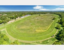 Luftaufnahme einer großen, ovalen Pferderennbahn mit umliegenden Wiesenflächen und begrünten Böschungen. Im Hintergrund sind vereinzelt Gebäude, Bäume und eine weite Landschaft unter klarem Himmel zu sehen.