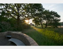 Eine geschwungene Sitzbank aus Ziegelstein und Metall steht in einer parkähnlichen Umgebung mit hohen Bäumen, dichtem Gebüsch und einer Wiese. Im Hintergrund ist eine kleine Backsteinkapelle sichtbar, die von der tief stehenden Sonne beleuchtet wird.