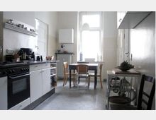 Helle Küche mit weiß gestrichenen Wänden und hexagonalem Fliesenboden, ausgestattet mit einer schwarzen Arbeitsplatte, Herd und weißen Unterschränken. Im Hintergrund steht ein Esstisch mit vier Stühlen vor einem großen Fenster, daneben befinden sich ein Heizkörper und ein weißer Wandkessel.