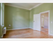 Leerer Innenraum mit hellgrün gestrichenen Wänden, hohen Decken mit weißer Stuckleiste und breiten Holzdielen. Links ein Fenster mit Heizkörper darunter, rechts eine weiße Kassettentür, die in einen weiteren Raum mit ähnlichem Holzboden führt.