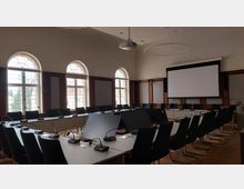 Konferenzraum mit langen, rechteckigen Tischen in U-Form, umgeben von schwarzen Stühlen. Der Raum hat hohe Fenster mit Rundbögen, eine Leinwand an der Stirnseite und moderne Mikrofone auf den Tischen.