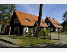 Traditionelle Fachwerkhäuser mit schwarzen Holzfassaden und roten Ziegeldächern, umgeben von Gartenanlagen und Straßenpflaster. Im Vordergrund ein Gehweg mit einer Haltestelle sowie ein schmiedeeisernes Tor, umgeben von Bäumen und Wiese.