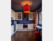 Eine gemütliche Küche mit cremefarbenen Schränken, dunkler Arbeitsfläche und einem Herd in der Mitte unter einer Dunstabzugshaube. Ein roter, hängender Lampenschirm beleuchtet den Raum, der einen Holzboden, eine lila Wandverkleidung und ein Fenster mit Vorhang zeigt; rechts stehen ein kleiner Tisch und gepolsterte Hocker.