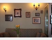 Wohnzimmer mit einer grün gemusterten Couch, einer Glasvase mit gelben Tulpen auf einem Holztisch und einer Wand mit mehreren historischen gerahmten Fotografien. Die Wand wird von zwei antiken Wandleuchtern beleuchtet, und ein verglastes Regal ist rechts im Bild sichtbar.