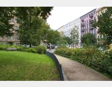 Grüne Parklandschaft mit einem Gehweg, der von Blumenbeeten und einem niedrigen Zaun begrenzt wird, umgeben von mehrstöckigen Wohnhäusern mit farbigen Fassaden im Hintergrund. Auf der linken Seite befindet sich eine Bank neben einem gepflegten Rasen und großen Bäumen.