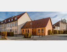 Ein kleiner Platz mit Kopfsteinpflaster, umgeben von Gebäuden mit roten Ziegeldächern, darunter ein orangefarbenes Haus mit Holztor und Sprossenfenstern. Im Hintergrund eine alte Ziegelmauer, Büsche und laternenartige Straßenbeleuchtung.