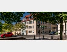 Gebäude mit heller Fassade und roten Dachziegeln an einer gepflasterten Straße, umgeben von Bäumen. Im Vordergrund stehen ein rotes und ein schwarzes Auto, daneben Straßenschilder und Fahrräder.