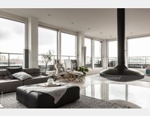 Modernes Wohnzimmer mit großzügigen Glasfronten, die auf eine Terrasse führen und viel Tageslicht hereinlassen. Der Raum ist mit einem Hängekamin, einer grauen Ledercouch, Sesseln sowie Pflanzen dekoriert und hat eine helle, minimalistische Einrichtung.