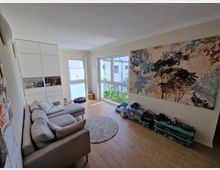 Wohnzimmer mit hellem Laminatboden, einer beige Couch und einem runden Teppich. Auf der linken Seite befindet sich ein weißer Einbauschrank, rechts ein großes Wandbild mit Baum-Motiv, und durch die Fenster fällt Tageslicht ein.