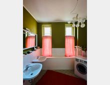 Badezimmer mit dunkelgrünen Wänden, weiß gefliester Badewanne und zwei kleinen Fenstern mit roten Vorhängen. Links hängen Spiegel und Waschbecken, rechts stehen eine Waschmaschine und eine Arbeitsfläche unter einer auffälligen Deckenleuchte mit kugelförmigen Lampen.