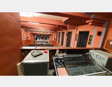 Blick aus einer Regiekabine in einen kleinen Theatersaal mit Bühnenbeleuchtung und Vorhängen an den Wänden. Im Vordergrund befinden sich ein Mischpult und technische Geräte, im Hintergrund ist die Bühne mit Requisiten zu sehen.