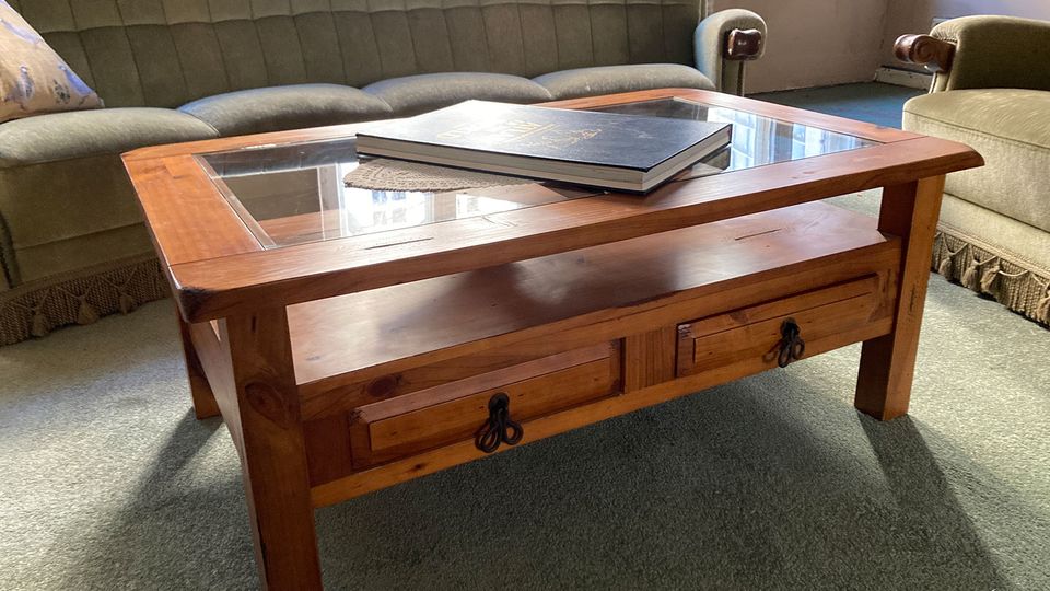 Wohnzimmer mit grün gepolsterter Couch und Sessel auf Teppichboden, davor ein rechteckiger Holzcouchtisch mit Glasplatte und zwei Schubladen. Auf dem Tisch liegt ein großes Buch, im Hintergrund ist ein Fensterbereich zu sehen.