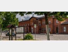 Backsteinbahnhof von Lübben mit einer Haltestellentafel im Vordergrund, umgeben von Kopfsteinpflaster, Bäumen und einem überdachten Informationsstand. Der Himmel ist teils bewölkt.