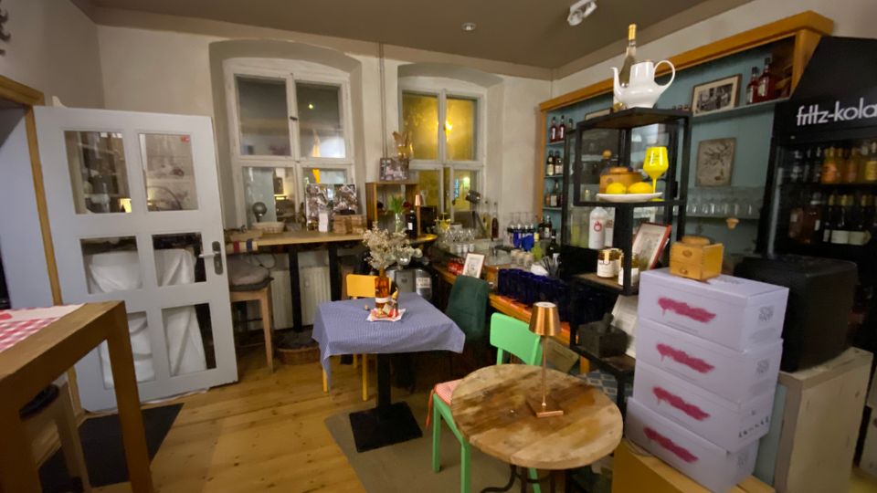 Gemütlicher Innenraum einer kleinen Bar oder eines Cafés mit Holzboden, kleinen Tischen und Stühlen sowie einer Theke mit Flaschenregalen. Im Hintergrund sind zwei Fenster, eine Glastür und ein Getränkekühlschrank zu sehen, daneben gestapelte Kisten und Dekoration auf Regalen.