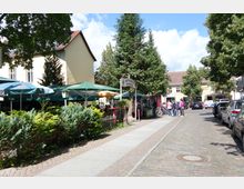 Straßenszene mit einem Biergarten auf der linken Seite, gekennzeichnet durch grüne Sonnenschirme und dichtes Gebüsch, sowie Fußgänger und geparkte Autos entlang eines gepflasterten Gehwegs und einer Straße. Im Hintergrund sind beige Gebäude mit roten Ziegeldächern und Bäumen zu sehen.