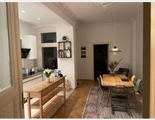 Helle Wohnküche mit integriertem Essbereich, ausgestattet mit weißen Küchenschränken, einer Arbeitsinsel aus Holz und einem hölzernen Esstisch mit bunten Stühlen. Stuckverzierungen an der Decke, ein Teppich unter dem Esstisch und eine moderne Pendelleuchte prägen den Raum.